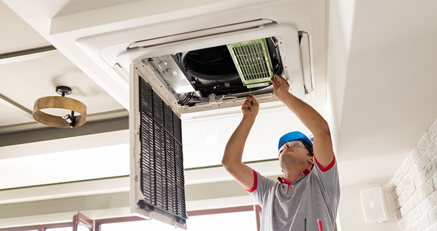 4 Things to Consider When Buying a New Central Air Unit for a Rental man on ladder installing air conditioning unit in ceiling opening