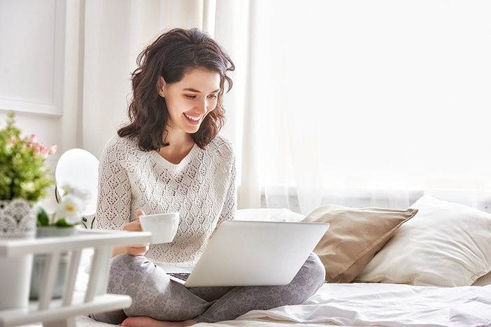 13 Simple Hacks to Dramatically Lower Your Healthcare & Childcare Costs Happy casual beautiful woman working on a laptop sitting on the bed in the house.