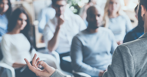 Newbies: Don’t Fall for This Real Estate Investing Scam! Rear view of man gesturing with hand while standing against defocused group of people sitting at the chairs in front of him