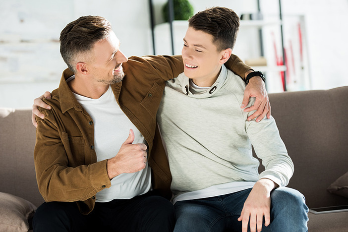 7 Ways to Screw Up Your Kid’s Financial Future (& How to Avoid It) smiling father and teen son hugging on sofa at home and looking at each other