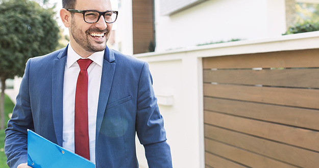 Should I Get My Real Estate License? An Examination of Benefits & Costs smiling brown-haired man with glasses and shadow of a beard evaluates exterior of a residential property while wearing suit and carrying a folder