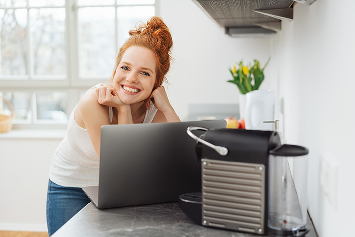 3 Habits of Incredibly Lucky People (For Better Fortune in Business & Life!) Young woman relaxing in a kitchen with a laptop computer balanced on the counter looking over the top to smile happily at camera