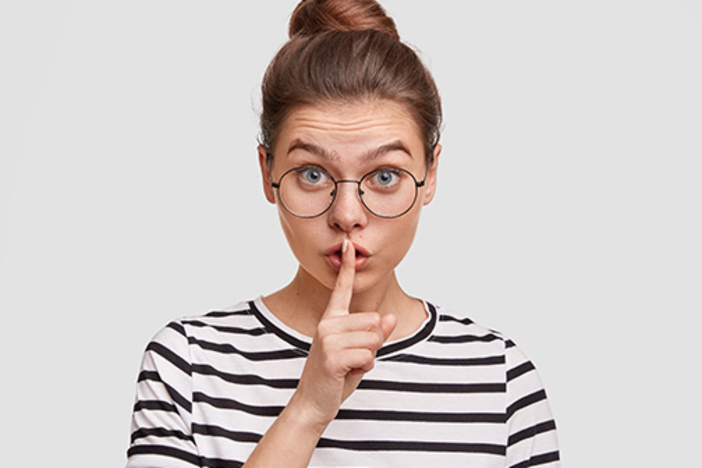 Nearly 25% of Americans Lie to Their Spouses About Money: It’s Time to Open Up About Finances Secret concept. Beautiful European female in eyewear, makes shush gesture, asks to keep voice down as suffers from headache, wears spectacles and striped t shirt, isolated over white background