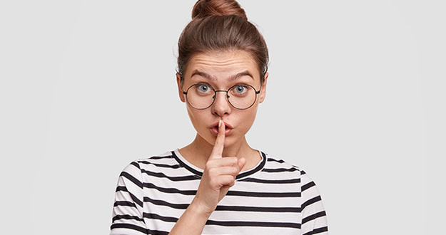 Nearly 25% of Americans Lie to Their Spouses About Money: It’s Time to Open Up About Finances Secret concept. Beautiful European female in eyewear, makes shush gesture, asks to keep voice down as suffers from headache, wears spectacles and striped t shirt, isolated over white background